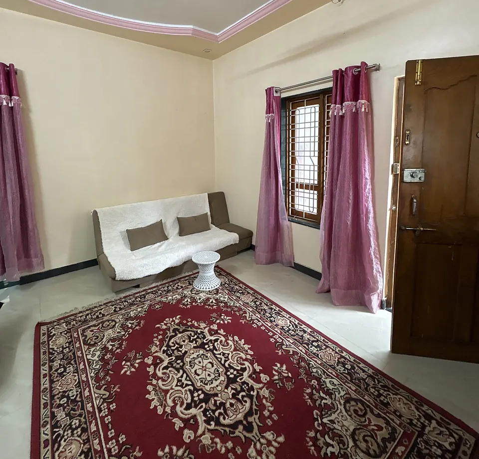 The hall seen from a corner, with the sofa-cum-bed under a window with pink curtains, the red carpet and the wooden front door