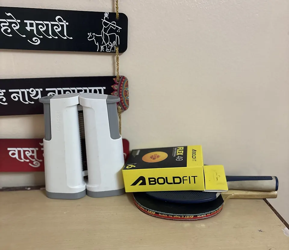 Portable table-tennis net, two bats and a box of balls below Devanagari name boards